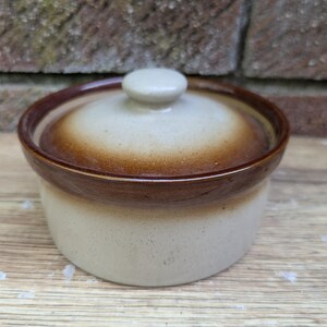 Puede incluir: Un pequeño recipiente redondo de cerámica con tapa, cuerpo beige moteado y borde y tapa marrón. La tapa tiene un pequeño pomo redondo. El recipiente está sobre una superficie de madera.