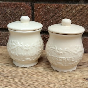 Könnte beinhalten: Zwei cremefarbene Keramikgläser mit Deckel. Die Gläser haben eine abgerundete Form mit geprägten floralen Designs und den Worten "Farm Aid" und "Reserve". Die Gläser stehen auf einer Holzoberfläche.