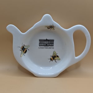 Op de afbeelding: Witte, keramische theepotvormige theezakjeshouder met een zwart-witte afbeelding van Chatsworth House en drie gele en zwarte bijen.