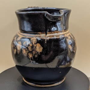 May include: A handmade, dark brown ceramic jug with a spout and handle. The jug has a glossy finish with areas of tan and brown marbling. The base is a darker shade of brown. The jug is sitting on a black surface.