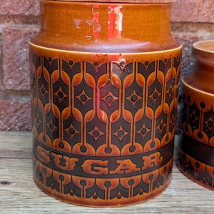 May include: A vintage brown ceramic sugar jar with a wooden lid. The jar features a repeating geometric pattern in dark brown and orange, with the word "SUGAR" in a bold font. The jar is cylindrical and has a glossy finish.