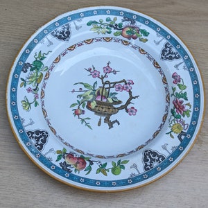 May include: A white ceramic plate with a blue and gold border. The center of the plate features a floral design with a basket of fruit and a bird.