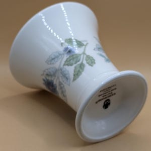 May include: White ceramic vase with a floral design featuring blue and green flowers and leaves. The vase has a flared top and a round base. The bottom of the vase is marked with the text "WEDGWOOD MADE IN ENGLAND"
