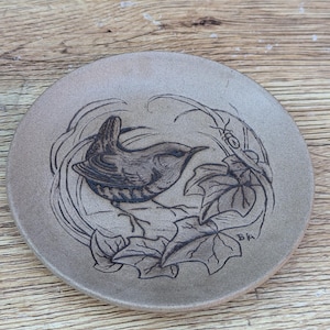 May include: A circular, tan ceramic plate featuring an etched design of a bird amidst leaves and vines. The bird is facing right, with detailed wings and body. The plate has a rustic, handmade appearance, with a diameter of approximately 15 cm.