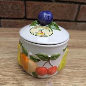 Pode incluir: Jarro de cerâmica branco com tampa que apresenta um desenho pintado à mão de fruta, incluindo peras, cerejas e uma laranja. A tampa tem uma ameixa azul e roxa no topo. O jarro tem um rótulo que diz "Country Kitchen by Rayware Hand Painted Ceramics".