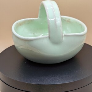 Può includere: Un piccolo cestino in ceramica verde chiaro con manico. Il cestino ha una forma arrotondata e una finitura liscia e lucida. Il manico è arcuato e attaccato ai lati del cestino. Il cestino è su una superficie nera.