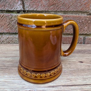 Puede incluir: Una jarra de cerveza de cerámica marrón con un asa redondeada y una banda decorativa alrededor de la base.