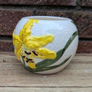 Op de afbeelding: Een witte keramische vaas met een getextureerd oppervlak. Aan de zijkant is een grote, gele leliebloem met bruine vlekken en groene bladeren geschilderd. De vaas heeft een ronde vorm en een brede opening.