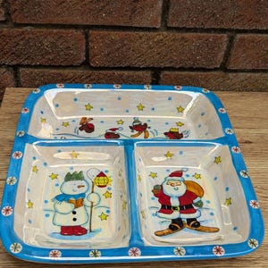 May include: A white ceramic serving dish with four sections. The dish is decorated with a winter holiday theme, featuring a snowman, Santa Claus, and snowflakes. The dish has a blue rim with white snowflakes.