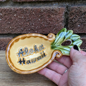 Puede incluir: Un plato de cerámica con forma de piña, centro amarillo y hojas verdes. El texto Aloha Hawaii está grabado en letras negras en el centro del plato. El plato tiene un acabado brillante y un borde festoneado alrededor de la parte amarilla.