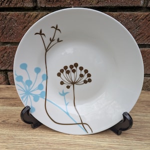 May include: White ceramic plate with a floral design in brown and light blue. The plate features a central flower and other floral elements. The plate is displayed on a black stand against a brick background.
