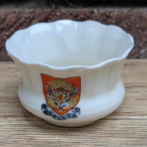 May include: Cream-colored ceramic bowl with a scalloped rim. The bowl features a colorful crest with the word "KELSO" on a banner. The crest includes a shield, trees, and birds. The bowl sits on a wooden surface.