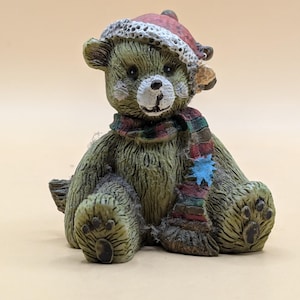 Pode incluir: Uma figura de cerâmica de um urso de peluche verde usando um chapéu de Papai Noel vermelho e branco e uma echarpe colorida. O urso tem um nariz branco e olhos pretos.