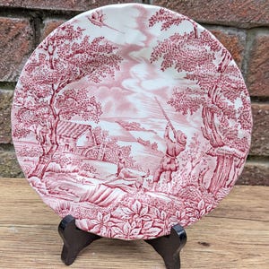 May include: A round plate with a red and white pastoral scene. The design features a hunter, a dog, trees, and a building. The plate is displayed on a black wooden stand, evoking a vintage aesthetic. The plate's diameter is approximately 20 cm.