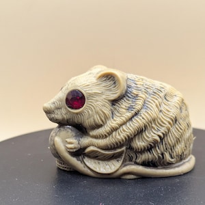 Puede incluir: Una detallada figura de ratón de color beige, tallada con pelaje texturizado y sosteniendo un objeto pequeño. El ratón tiene un prominente ojo de gema roja. La figura descansa sobre una superficie negra.