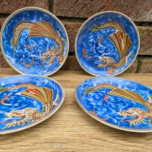 May include: Four blue ceramic dishes with a dragon design. The dishes have a white rim and a vibrant blue background with a swirling pattern. Each dish features a detailed dragon in gold, red, and orange.