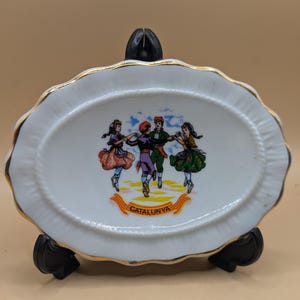 May include: White oval ceramic dish with a gold rim and a scene of four people dancing in traditional Catalan clothing. The dish has the word "CATALUNYA" written in gold lettering.