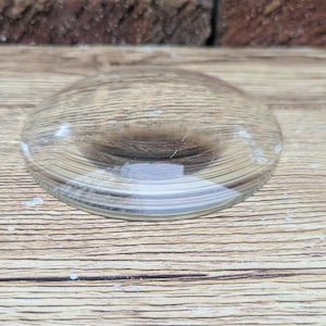 May include: A clear, glass lens with a rounded shape. The lens is slightly raised in the center and has a smooth, transparent surface. The lens is set on a wooden surface.