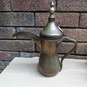 Può includere: Una caffettiera araba in ottone con un beccuccio e un manico. La caffettiera ha un coperchio decorativo con un pomo.