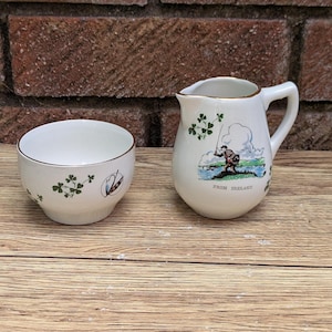 May include: A white ceramic creamer and sugar bowl set. The creamer features a handle and a painted scene of a fisherman with the text "FROM IRELAND". The sugar bowl and creamer are decorated with green shamrocks and gold trim.