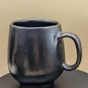 Könnte beinhalten: Eine schwarze Keramiktasse mit rundem Körper und gebogenem Henkel. Die Tasse hat eine matte Oberfläche und steht auf einer schwarzen Oberfläche. Die Tasse ist für Heißgetränke konzipiert.