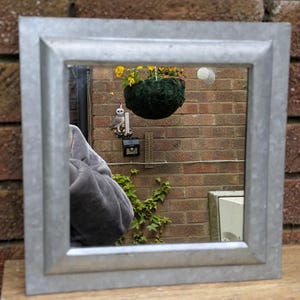 May include: A square mirror featuring a wide, flat, metallic frame with a galvanised steel finish. The reflective surface shows a clear view of an outdoor brick wall, a hanging plant with yellow flowers, a small owl figurine, and a thermometer.