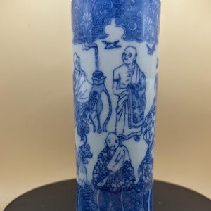 May include: A cylindrical blue and white porcelain vase. The vase features a detailed design of figures and floral patterns. The top and bottom rims have a blue floral border. The figures are dressed in robes. The vase is on a black surface.