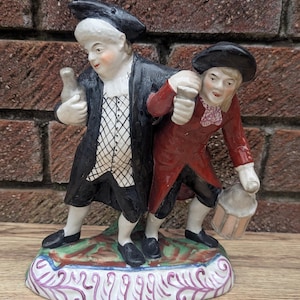 Peut inclure: Une figurine en porcelaine représentant deux personnes, l'une portant un manteau et un chapeau noirs, tenant une bouteille, et l'autre portant un manteau et un chapeau rouges, tenant une tasse. La figurine est posée sur un socle coloré avec un motif floral.