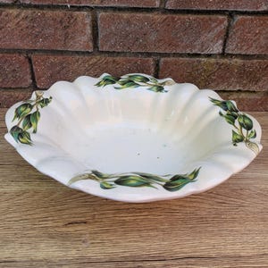 Puede incluir: Bol de cerámica blanco con borde festoneado y diseño de hojas verdes. El cuenco tiene una forma ancha y poco profunda y está decorado con un patrón repetido de hojas y tallos verdes alrededor del borde. El cuenco está sobre una superficie de madera.