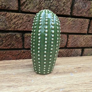May include: A green ceramic cactus-shaped vase with white dot details. The vase is cylindrical and stands upright on a wooden surface. The background features a red brick wall.