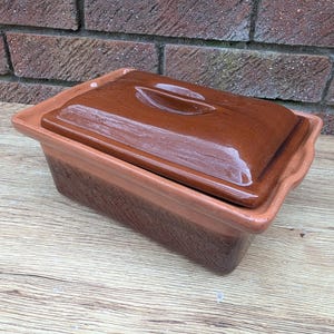 May include: A rectangular, brown ceramic baking dish with a matching lid. The dish has a textured exterior and a glossy, smooth lid with a small handle. The dish is designed for baking and serving.