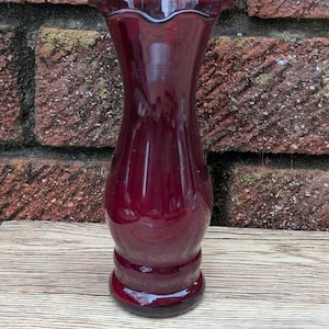 Può includere: Un vaso di vetro rosso scuro con bordo increspato. Il vaso ha una base bulbosa e un corpo sottile e allungato. Si trova su una superficie di legno contro un muro di mattoni. Il vaso è alto circa 20 cm.