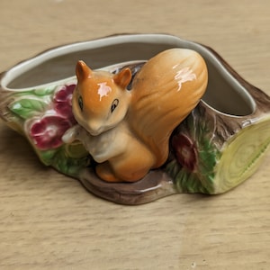 Puede incluir: Una maceta de cerámica con forma de tronco con una detallada figura de ardilla. La ardilla es naranja y blanca, con una cola tupida. La maceta tiene un exterior marrón, un interior crema y está decorada con detalles florales rojos y verdes.