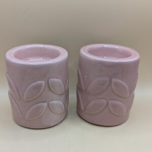 Op de afbeelding: Twee roze keramische kaarshouders met een bladontwerp.
