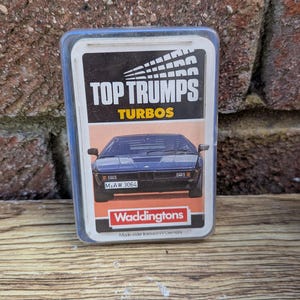 Puede incluir: Un juego de cartas Top Trumps Turbos vintage en una funda de plástico transparente. La carta presenta un coche deportivo azul oscuro y el texto "TOP TRUMPS TURBOS". El logotipo de Waddingtons es visible en la parte inferior.
