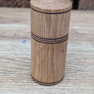 以下が含まれることがあります： 取り外し可能な蓋が付いた円筒形の木製容器。明るい茶色の自然な木目模様が特徴で、本体と蓋の上端を囲むように濃い茶色の水平なエッチングラインが施されており、シンプルで機能的なデザインに仕上がっています。