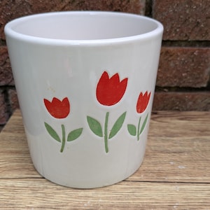 May include: White ceramic flower pot with a tulip design. The pot features three red tulips with green stems and leaves. The pot is cylindrical and sits on a wooden surface.
