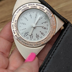 Puede incluir: Un reloj plateado con hora mundial, esfera blanca y números negros. El bisel muestra nombres de ciudades como Londres, Tokio y Nueva York. El reloj está en un estuche de cuero negro.
