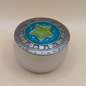 Könnte beinhalten: Eine kleine, runde, weiße Schmuckdose mit einem silbernen Deckel. Der Deckel hat ein blaues und grünes Emaille-Design mit einem fünfzackigen Stern in der Mitte.