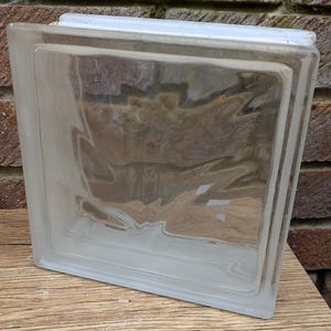 May include: A clear glass block with a rectangular shape and a frosted appearance. The block has a white top and is sitting on a wooden surface. The background features a brick wall.