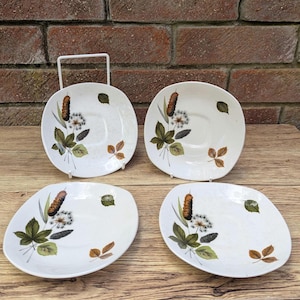 Peut inclure: Quatre assiettes en céramique blanche avec un motif inspiré de la nature. Chaque assiette présente un roseau, un pissenlit et des feuilles dans des tons de vert, de marron et de blanc. Les assiettes sont présentées sur une surface en bois.