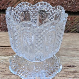 Op de afbeelding: Transparante glazen voetkom met een decoratief bloemen- en ruitpatroon. De kom heeft een geschulpte rand en staat op een ronde, geschulpte basis. Het glas is transparant. Waarschijnlijk een vintage object.