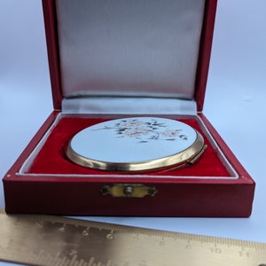 May include: A vintage compact mirror with a floral design in a red velvet box. The mirror is round and has a gold rim. The floral design is in shades of pink and green.