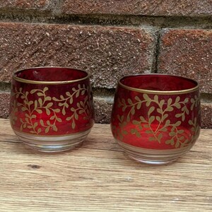 May include: Two red glass votive candle holders with gold leaf design. The candle holders have a clear base and a gold rim. The gold leaf design is a repeating pattern of leaves and vines. The candle holders are sitting on a wooden surface.