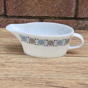 Puede incluir: Salsera de cerámica blanca con asa y pico vertedor. La salsera presenta una banda decorativa de motivos de copos de nieve azules y marrones alrededor del borde. Colocada sobre una superficie de madera.