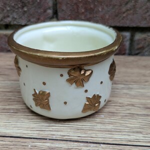 May include: A small, off-white ceramic pot with a gold rim and gold leaf and dot accents. The pot is round with a slightly flared top and sits on a wooden surface. The background is a brick wall.