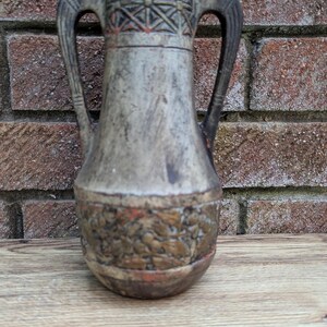 May include: A weathered, gray-toned ceramic vase with two handles. The top features a geometric pattern in red and brown, while the base has a textured, embossed design. The vase is set against a brick wall and wooden surface.