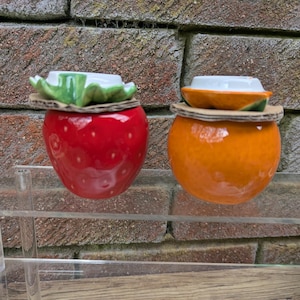 Könnte beinhalten: Zwei keramikförmige Obstbehälter. Einer ist eine glänzend rote Erdbeere mit einem grün-weißen blattförmigen Deckel. Der andere ist ein orangefarbener Behälter mit einem weiß-grünen Deckel. Beide stehen auf Kartonbasen.