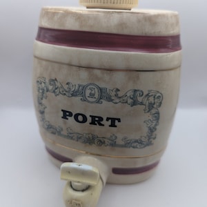May include: Cream-colored ceramic barrel-shaped container with a spigot and a removable lid. The container has a dark red band and the word "PORT" in black letters. Decorative blue scrollwork surrounds the word.