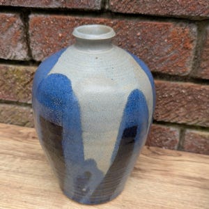 May include: A handcrafted ceramic vase with a rounded form and a slender neck. The vase displays a palette of blue, grey, and black, with blue abstract patterns against the grey. The vase is placed on a wooden surface.
